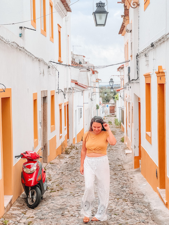 brunette in a yellow shirt and white pants standing in a street lined with white houses that have yellow window linings and a red motorcycle