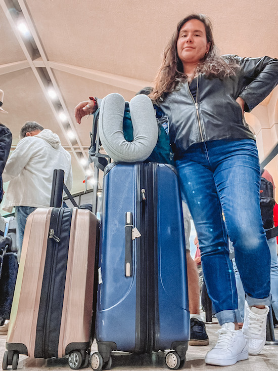 brunette woman wearing a leather jacket and blue jeans standing next to a blue large suitcase with a neck pillow and light pink carry on suitcase Moving Abroad Costs US to Portugal