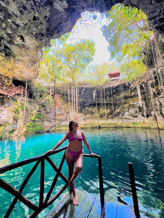 brunette standing in a cenote in Mexico that has blueish green water and an opening above with trees and the sky - Mexican Caribbean Coast