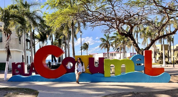 brunette sitting in front of a colored sign that says Isla Cozumel - Mexican Caribbean Coast