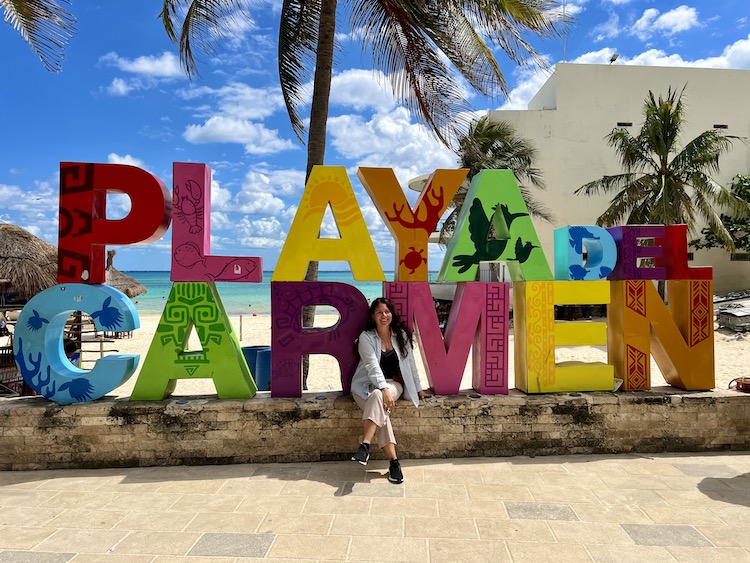 brunette woman sitting in front of the Playa del Carmen sign on the beach on a sunny day